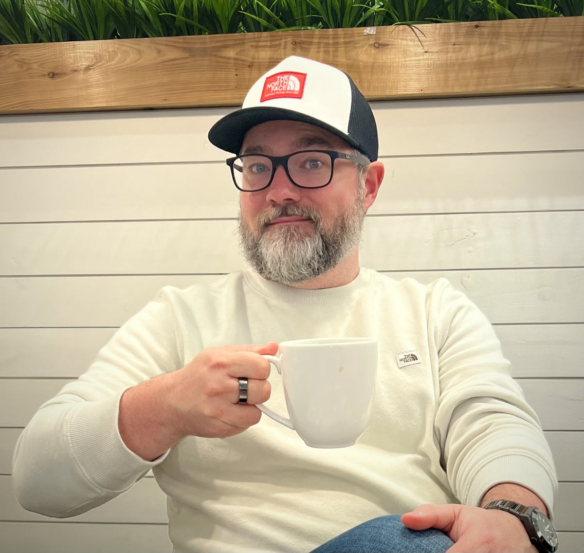 Our Founder, TJ Sloan. He is a man with a beard and glasses wearing a white and black trucker hat with a red The North Face logo, holding a white mug. He is wearing a cream-colored long-sleeve shirt and blue jeans, seated in front of a white wood-paneled wall with a wooden border and green plants.